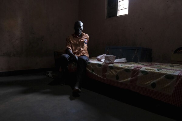 Simon Tigawalana sits on a bed as he rests his feet on a clay-based earthen floor made by EarthEnable at home on May 16, 2025, in Jinja, Uganda. (AP Photo/Hajarah Nalwadda)